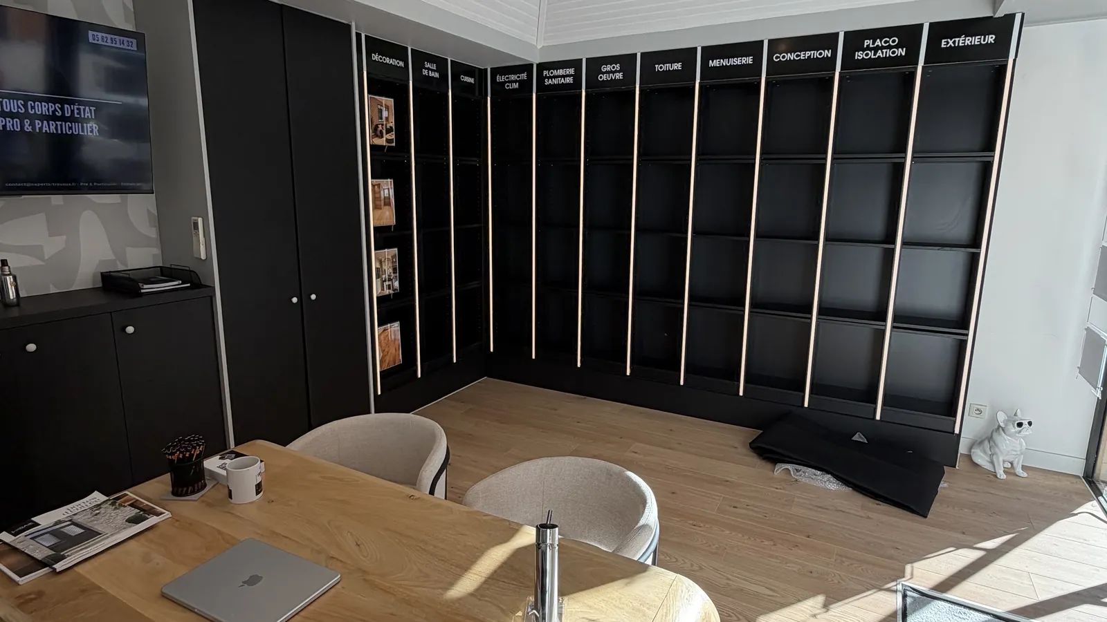 Accueil du showroom : un bureau en bois clair entouré de fauteuils, devant un mur de rangements noirs aux rubriques des métiers du bâtiment, sous un plafond en lambris blanc, sur un parquet clair.